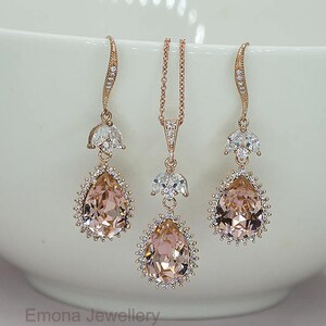 May include: A rose gold bridal jewelry set with a teardrop pendant necklace and matching earrings. The pendant and earrings feature a large blush pink crystal surrounded by smaller clear crystals.