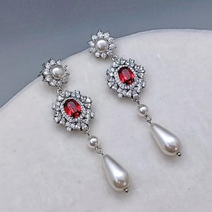 May include: Elegant silver earrings featuring a central oval red gemstone surrounded by sparkling clear crystals. Each earring has a pearl accent and a dangling teardrop-shaped pearl. The earrings are designed for a formal occasion.