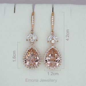 May include: Rose gold dangle earrings with a pear-shaped blush pink crystal surrounded by smaller clear crystals. The earrings measure 4.2 cm in length and 1.2 cm in width.