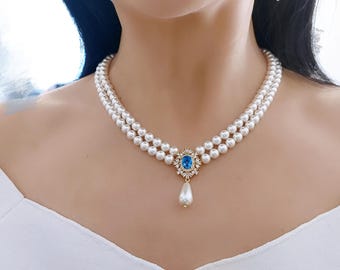 Puede incluir: Un collar de perlas de doble hebra con un colgante de piedra azul. El colgante está rodeado de pequeñas piedras transparentes y tiene una gota de perla en forma de pera. El collar se lleva sobre una parte superior blanca.