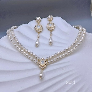 Peut inclure: Un ensemble collier et boucles d'oreilles dorées. Le collier est composé de deux rangées de perles blanches et d'un pendentif central orné d'une pierre ovale. Les boucles d'oreilles reprennent le même design avec une perle pendante.