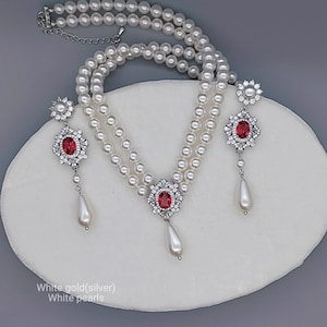 May include: A white pearl necklace and earring set. The necklace has three strands of pearls and a silver clasp. The earrings and necklace pendant feature a red gemstone surrounded by clear crystals. The text in the image says "White gold(silver) White pearls."