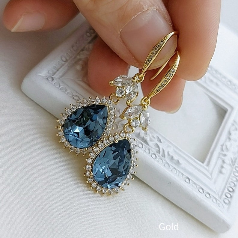 Op de afbeelding: Gouden oorhangers met een traanvormige blauwe kristal omringd door kleinere heldere kristallen. De oorbellen hebben een bloemmotief met heldere kristallen.