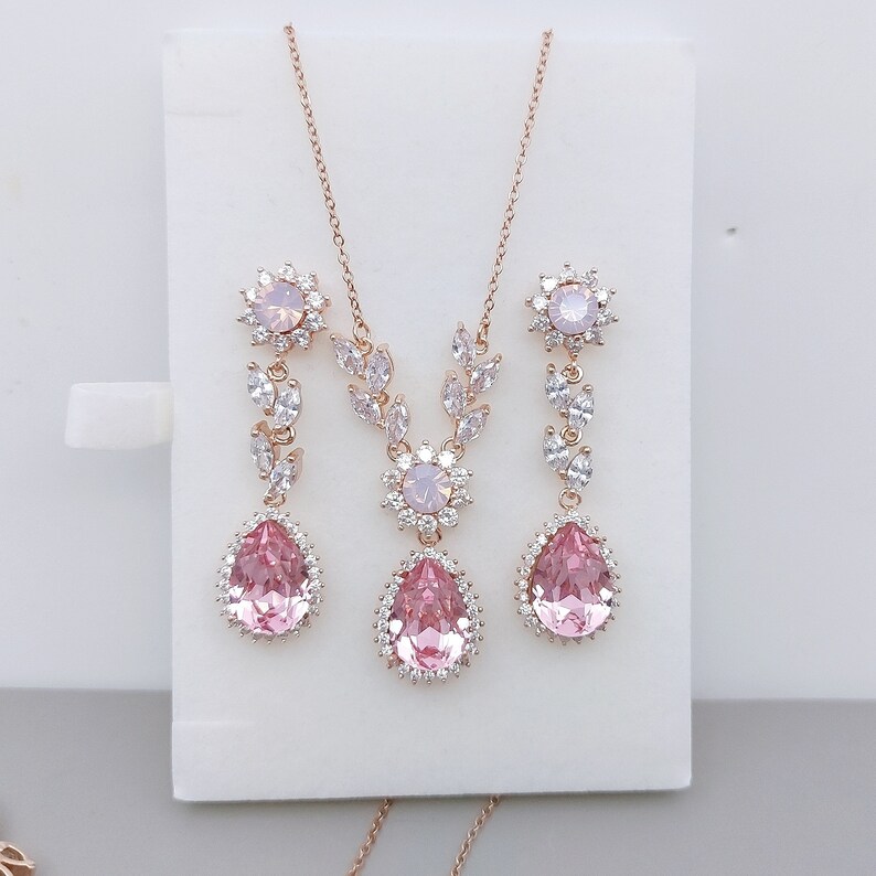 Puede incluir: Un collar y pendientes de oro rosa con detalles de cristal rosa y blanco. El collar presenta un delicado dise&ntilde;o floral con un colgante en forma de l&aacute;grima. Los pendientes tienen un dise&ntilde;o similar con un colgante en forma de l&aacute;grima m&aacute;s peque&ntilde;o.