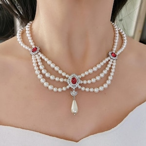 May include: A three-strand pearl choker necklace with a silver pendant featuring a red gemstone surrounded by smaller clear gemstones. The pendant is attached to a single pearl drop.