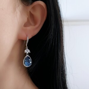 Op de afbeelding: Een paar zilveren oorhangers met een traanvormige blauwe edelsteen omringd door kleinere heldere edelstenen. De oorbellen hebben een delicaat bloemenmotief.