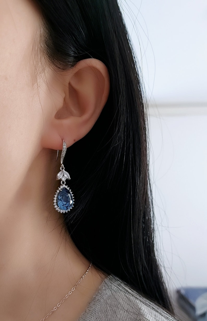 Op de afbeelding: Een paar zilveren oorhangers met een traanvormige blauwe edelsteen omringd door kleinere heldere edelstenen. De oorbellen hebben een delicaat bloemenmotief.