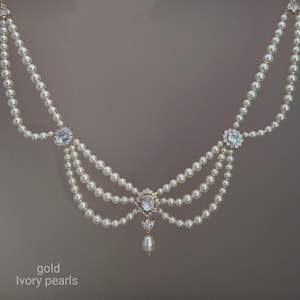 Può includere: Una collana a più fili con perle color avorio disposte in un design drappeggiato. La collana presenta tre montature centrali in metallo dorato incastonate con cristalli trasparenti e un pendente a goccia di perla al centro.
