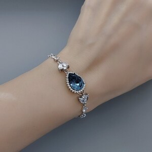 Op de afbeelding: Een zilveren armband met een grote, traanvormige blauwe kristal omringd door kleinere, heldere kristallen.