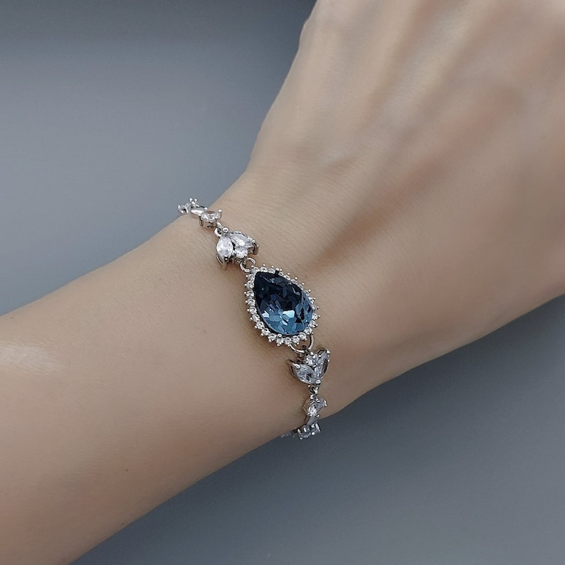Op de afbeelding: Een zilveren armband met een grote, traanvormige blauwe kristal omringd door kleinere, heldere kristallen.