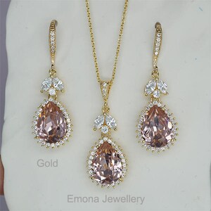 May include: A gold necklace and earrings set with a pear-shaped pink crystal pendant and matching earrings. The pendant and earrings are surrounded by smaller clear crystals.