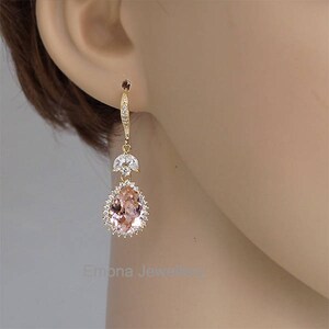 May include: A pair of gold dangle earrings with a teardrop-shaped blush pink crystal surrounded by smaller clear crystals.