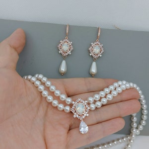 Op de afbeelding: Een twee-strengs parelketting met een roosgoud hanger met een grote ovale, heldere steen omgeven door kleinere heldere stenen. Passende roosgoud oorbellen met een heldere steen en kleinere heldere stenen hangen aan een parel.