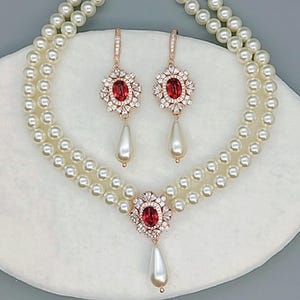 Op de afbeelding: Een parelketting en oorbellen set. De ketting heeft twee strengen witte parels. De oorbellen en de hanger van de ketting hebben een rode ovale steen omringd door heldere kristallen en een bungelende parel. Het metaal is roségoud.