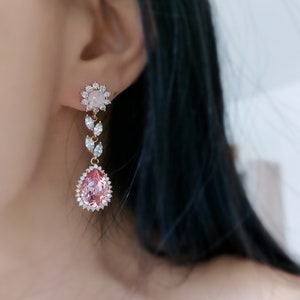 Puede incluir: Un par de pendientes colgantes con una piedra preciosa rosa en forma de pera rodeada de cristales transparentes. Los pendientes tienen una piedra preciosa redonda blanca m&aacute;s peque&ntilde;a rodeada de cristales transparentes en la parte superior.