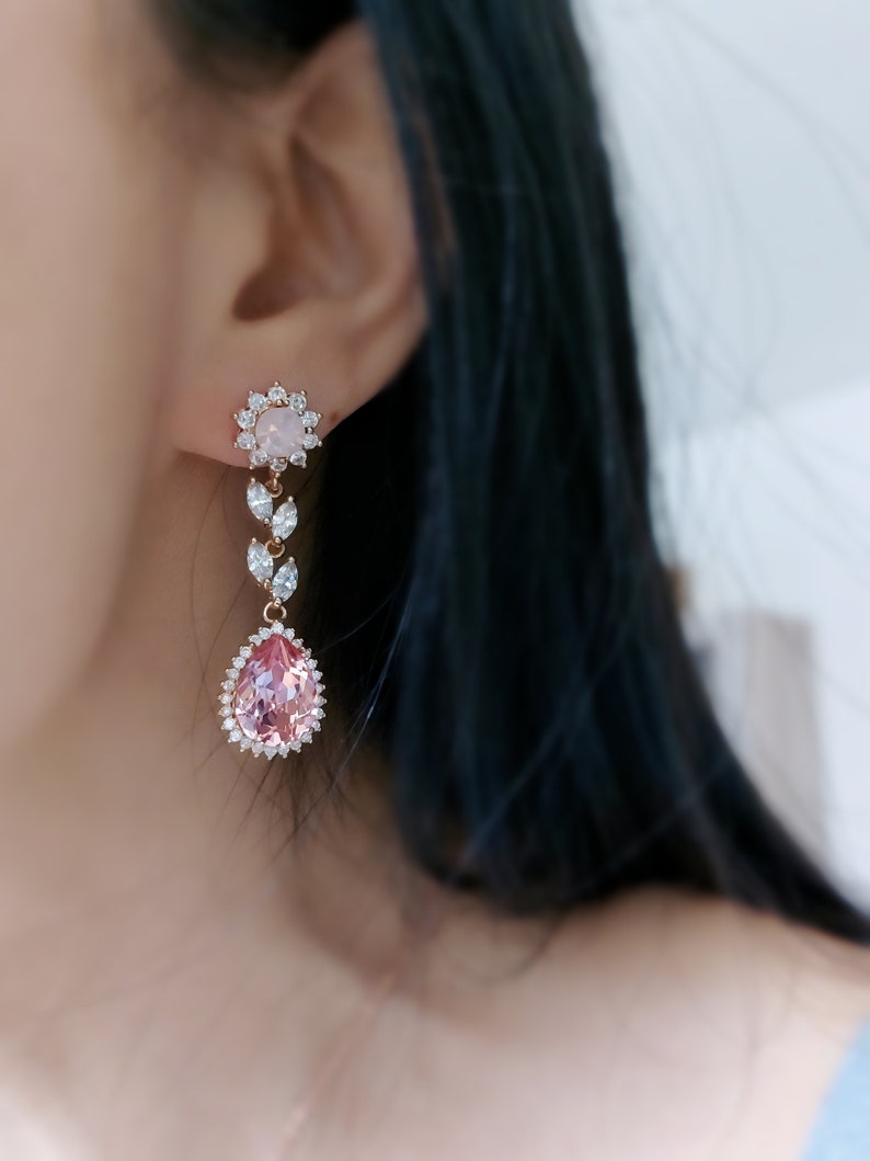 Puede incluir: Un par de pendientes colgantes con una piedra preciosa rosa en forma de pera rodeada de cristales transparentes. Los pendientes tienen una piedra preciosa redonda blanca m&aacute;s peque&ntilde;a rodeada de cristales transparentes en la parte superior.