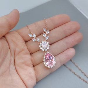 Puede incluir: Un delicado collar de oro rosa con un gran colgante de cristal rosa en forma de l&aacute;grima rodeado de cristales transparentes m&aacute;s peque&ntilde;os. El collar presenta un peque&ntilde;o grupo de cristales transparentes sobre el colgante.