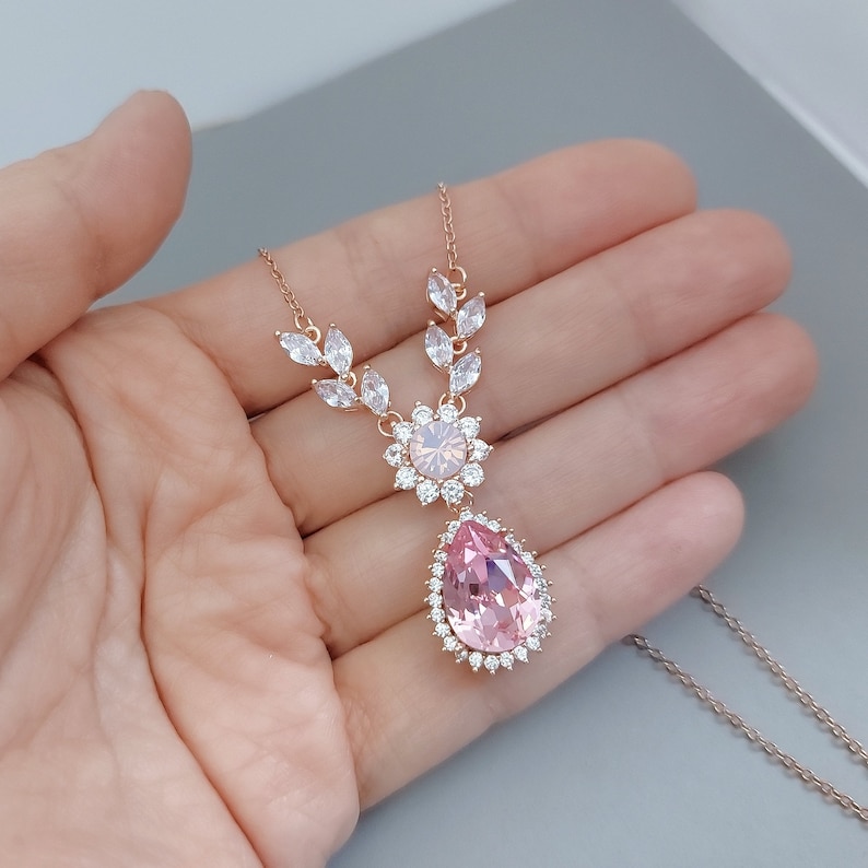 Puede incluir: Un delicado collar de oro rosa con un gran colgante de cristal rosa en forma de l&aacute;grima rodeado de cristales transparentes m&aacute;s peque&ntilde;os. El collar presenta un peque&ntilde;o grupo de cristales transparentes sobre el colgante.