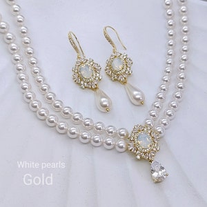 Può includere: Una collana di perle a doppio filo con un ciondolo dorato che presenta una grande pietra trasparente circondata da pietre trasparenti più piccole. Orecchini dorati abbinati con pietre trasparenti che pendono dalla parte inferiore.