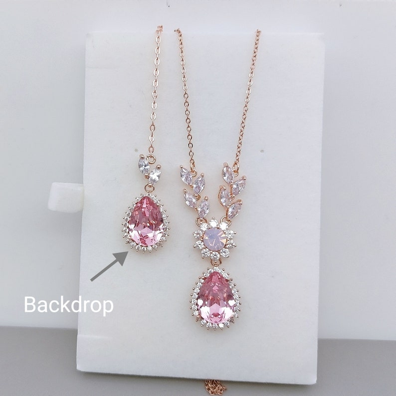Puede incluir: Un conjunto de collar y pendientes de oro rosa con un colgante de cristal rosa en forma de l&aacute;grima y un dise&ntilde;o floral. El collar tiene una cadena delicada y los pendientes son de estilo gota.