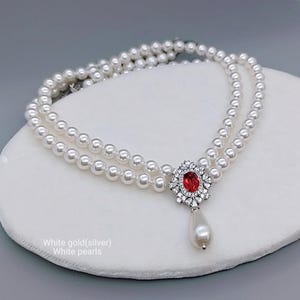 Può includere: Una collana di perle a due fili con un pendente con una gemma rossa circondata da cristalli trasparenti. Una singola perla a goccia pende sotto. Il testo "White gold(silver) White pearls" è visibile.
