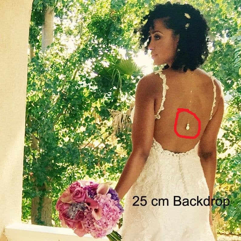 Puede incluir: Una novia con un vestido de novia de encaje blanco con la espalda descubierta. El vestido tiene una delicada cadena dorada con un peque&ntilde;o colgante que cuelga de la parte posterior del cuello. La novia lleva un ramo de flores rosas y moradas. El texto "25 cm Backdrop" es visible en la esquina inferior derecha de la imagen.