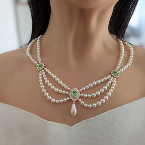 Può includere: Una collana di perle a più fili con tre file drappeggiate di perle bianche, arricchita da tre pietre preziose ovali verdi incastonate in metallo dorato e un singolo ciondolo di perla a goccia appeso alla montatura centrale.