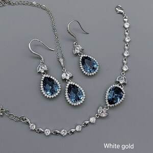 Op de afbeelding: Een set met druppelvormige oorbellen, een ketting en een armband. Elk stuk heeft een grote blauwe edelsteen omringd door kleinere heldere stenen. De sieraden zijn gemaakt van witgoud.