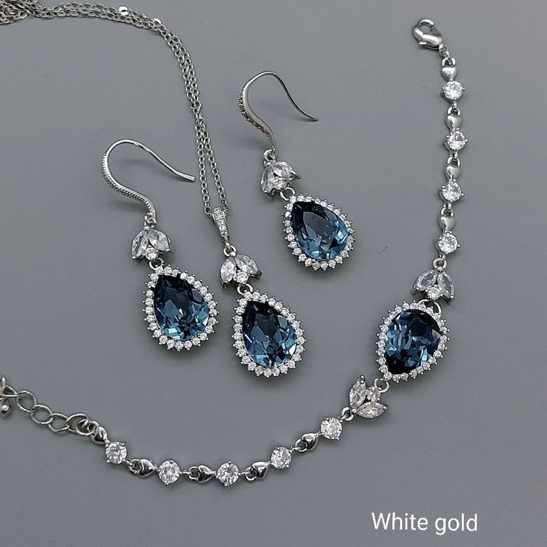 Op de afbeelding: Een set met druppelvormige oorbellen, een ketting en een armband. Elk stuk heeft een grote blauwe edelsteen omringd door kleinere heldere stenen. De sieraden zijn gemaakt van witgoud.