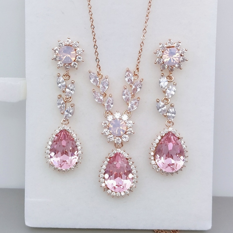 Puede incluir: Un conjunto de collar y aretes de oro rosa con cristales rosados en forma de pera y acentos de circonita c&uacute;bica transparentes. El collar presenta un delicado dise&ntilde;o de hojas y los aretes tienen un cristal en forma de pera colgante.