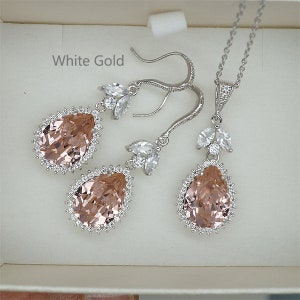 May include: A bridal jewelry set in white gold featuring a pear-shaped blush pink crystal surrounded by smaller clear crystals. The set includes a necklace and a pair of dangle earrings.