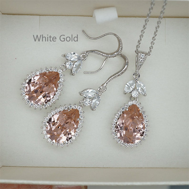 May include: A bridal jewelry set in white gold featuring a pear-shaped blush pink crystal surrounded by smaller clear crystals. The set includes a necklace and a pair of dangle earrings.