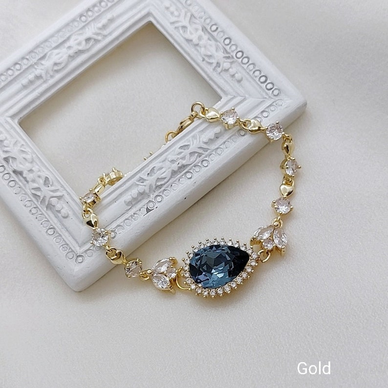 Op de afbeelding: Een gouden armband met een grote blauwe druppelvormige kristal omringd door kleinere heldere kristallen. De armband ligt op een wit oppervlak met een wit frame.