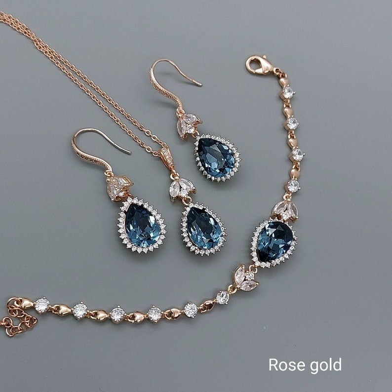 Op de afbeelding: Roze gouden sieradenset met een ketting, armband en oorbellen. Elk stuk heeft een traanvormige blauwe steen omringd door kleinere heldere stenen.