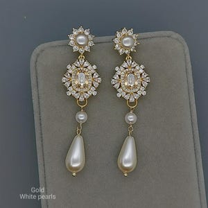 Op de afbeelding: Gouden oorhangers met witte parels. Elke oorbel heeft een bloemvormige bovenkant met een centrale parel, een diamantvormig midden, een kleine parel en een peervormige pareldruppel. De tekst "Gold White pearls" is zichtbaar.