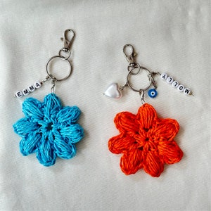 May include: Two crocheted flower keychains, one blue and one orange, with silver key rings and charms. The blue keychain has the word "EMMA" spelled out in white beads, and the orange keychain has the word "MOLLY" spelled out in white beads.