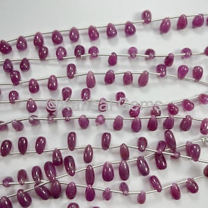 May include: Close-up of several strands of ruby beads. The beads are a deep pinkish-red hue, shaped like teardrops, and strung on thin, silver-coloured wire. The beads vary in size and shape, giving a natural, organic appearance.