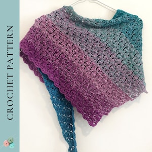 Puede incluir: Un chal de ganchillo en tonos de morado, turquesa y azul. El chal está hecho con un delicado patrón de punto y tiene un borde festoneado.