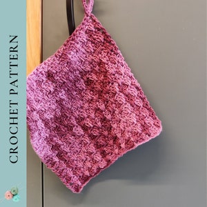 May include: A pink and purple crocheted dishcloth hanging on a hook. The dishcloth is made with a textured stitch and has a loop at the top for hanging.