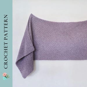 May include: A soft, purple crochet blanket draped over a white surface. The blanket is made with a simple stitch pattern and has a slightly textured look.