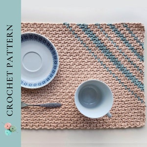 Op de afbeelding: Een gehaakte placemat in lichtbruin met een diagonaal ontwerp in blauwgroen. Een wit bord met een blauw patroon, een zilveren vork en een witte beker liggen op de placemat. De tekst "CROCHET PATTERN" staat links.