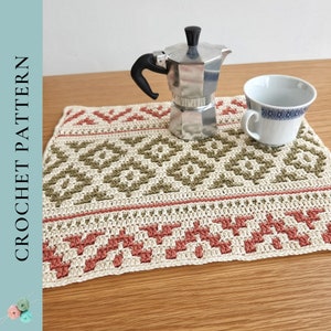 May include: A crocheted placemat with a geometric pattern in shades of beige, green, and red. The placemat is on a wooden table with a silver coffee pot and a white cup.