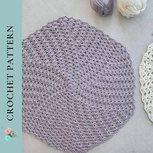 May include: A close-up of a crocheted doily in a light purple colour. The doily is round and has a delicate, lacy design.