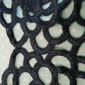 May include: Close-up of navy blue lace trim with a repeating circular pattern. The lace is stitched with a textured thread, creating a raised effect. The background is a light blue fabric with small white polka dots.
