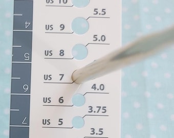 May include: A white and gray knitting needle gauge with numbered holes and US sizes from 0 to 15. The gauge also has millimeter measurements. A knitting needle is inserted into the gauge.