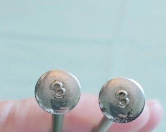 May include: Two silver-colored knitting needles with rounded tops. Each top has the number "3" etched into it. The needles are held against a light green background.