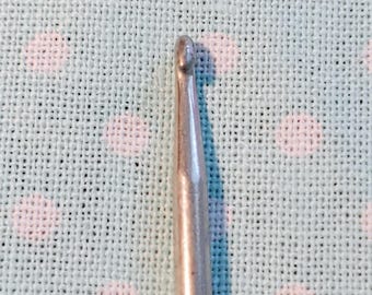 May include: Close-up of a silver crochet hook. The hook has a rounded tip and a smooth, metallic finish. The background is a light blue fabric with a white polka dot pattern.