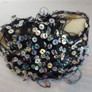 Puede incluir: Una bobina de hilo negro con lentejuelas iridiscentes adheridas. Las lentejuelas están dispuestas en un patrón aleatorio y son una mezcla de colores plata e arcoíris.