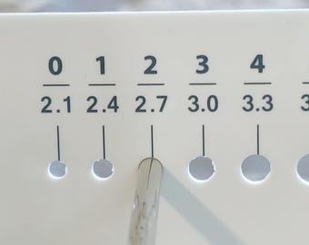 May include: A white plastic gauge with black numbers and measurements from 0 to 4, with corresponding millimeter measurements below. A clear, pointed tool is inserted into the hole labeled "2".