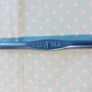 May include: A close-up of a blue metal crochet hook with the text "SIZE J USA" on the handle.